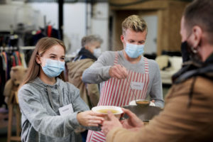 A young man and young woman volunteering at a soup kitchen, serving soup to a man in a brown jacket.